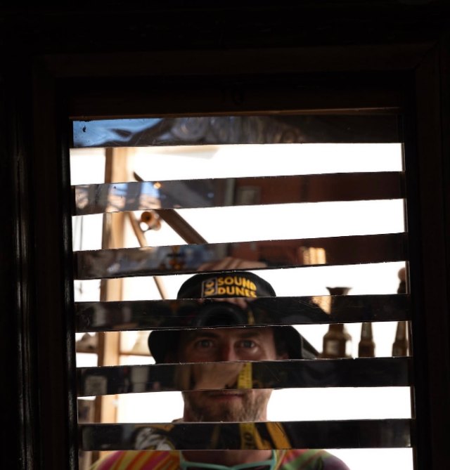 A person in a Sound Dunes cap looking through the slats of a window shutter in daylight