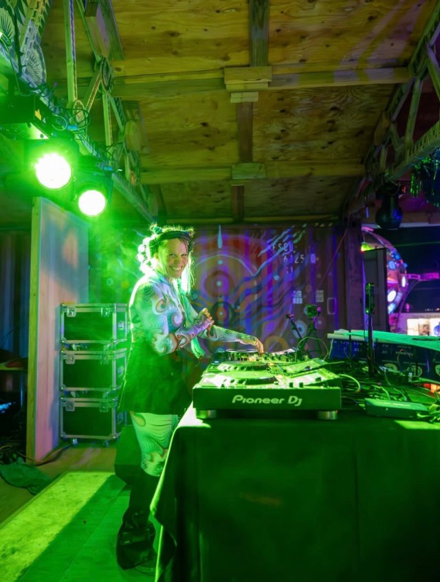 A DJ grinning behind Pioneer decks, lit green from a stage light in a timber-framed booth