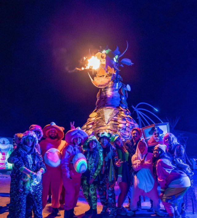 A group in bright costumes and onesies posing at night in front of a tall metal sculpture breathing fire